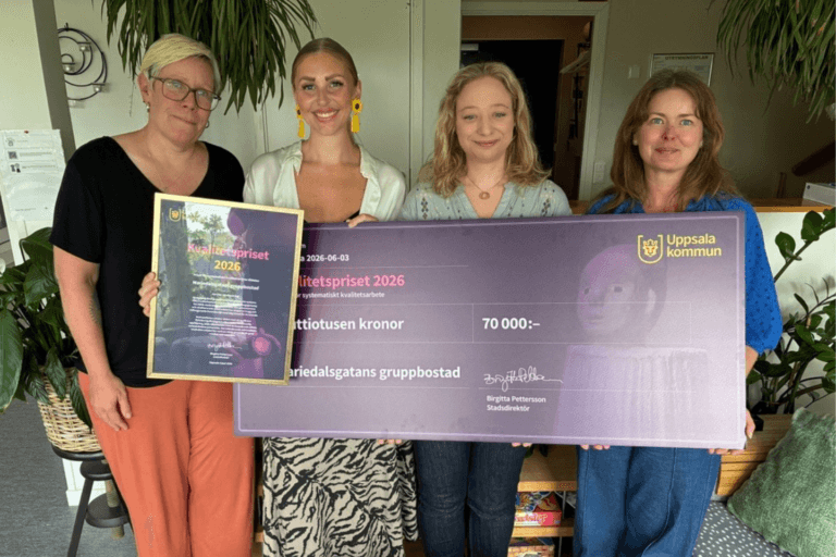 Fyra personer står inomhus och håller upp ett diplom och en stor prischeck från Uppsala kommun för Kvalitetspriset 2026. Checken är på 70 000 kronor och gäller Mariedalsgatans gruppbostad.