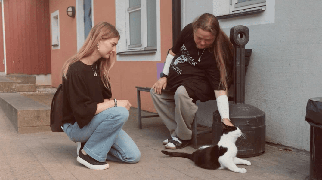 Två personer umgås med en svartvit katt utanför ett boende. En person sitter på huk och tittar på katten, medan den andra sitter på en bänk och klappar den. I bakgrunden syns en fasad med fönster och dörrar.