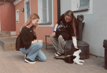 Två personer umgås med en svartvit katt utanför ett boende. En person sitter på huk och tittar på katten, medan den andra sitter på en bänk och klappar den. I bakgrunden syns en fasad med fönster och dörrar.