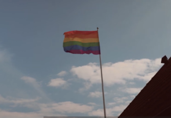 Regnbågsflagga vajande på en hög flaggstång mot en ljusblå himmel med vita moln. I bildens nedre högra hörn syns en del av ett mörkrött tak.