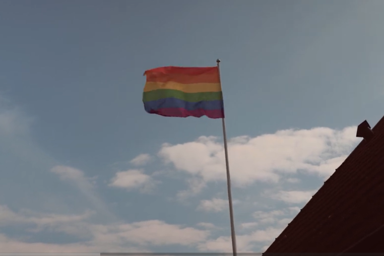 Regnbågsflagga vajande på en hög flaggstång mot en ljusblå himmel med vita moln. I bildens nedre högra hörn syns en del av ett mörkrött tak.