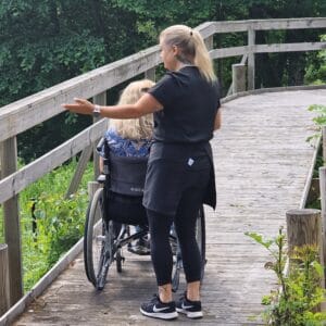 Person som står bredvid en rullstol på en bred träspång genom ett grönskande naturområde och pekar ut över omgivningen.