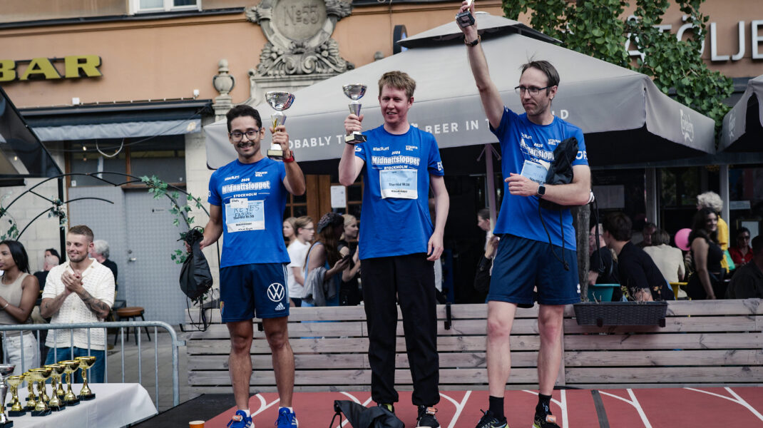 Tre deltagare prisas på en prispall efter Midnattsloppet och håller upp varsin pokal framför publik och uteserveringar i stadsmiljö.