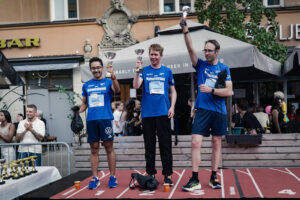 Tre deltagare prisas på en prispall efter Midnattsloppet och håller upp varsin pokal framför publik och uteserveringar i stadsmiljö.