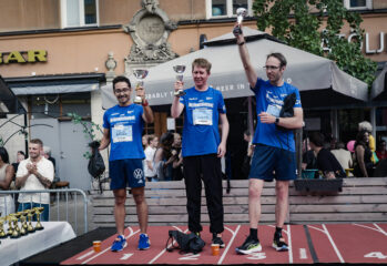 Tre deltagare prisas på en prispall efter Midnattsloppet och håller upp varsin pokal framför publik och uteserveringar i stadsmiljö.