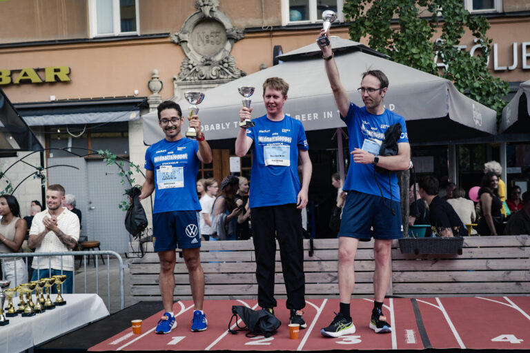 Tre deltagare prisas på en prispall efter Midnattsloppet och håller upp varsin pokal framför publik och uteserveringar i stadsmiljö.