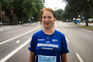 En deltagare i Midnattsloppet Stockholm 2026 poserar på tävlingsbanan i blå lopptröja och nummerlapp. Den breda stadsgatan omges av trädalléer och sträcker sig bakom personen i båda riktningarna.