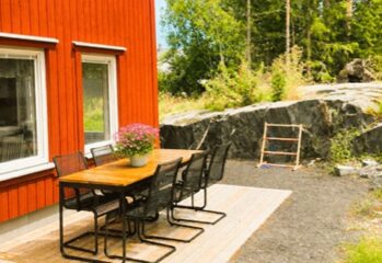 Uteplats med bord och stolar vid röd byggnad, med berg och grönska i bakgrunden.