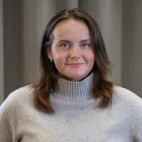 Porträttbild av en person med axellångt brunt hår och ljus stickad polotröja mot grå bakgrund.